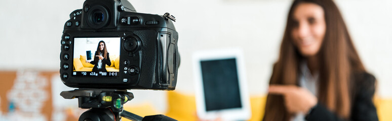 panoramic shot of digital camera with happy video blogger pointing with finger at digital tablet with blank screen