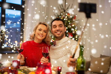 Obraz na płótnie Canvas christmas, holidays, technology and people concept - happy couple in taking picture by selfie stick at home dinner over snow