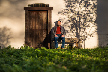 Lonely senior man sitting on bench in front of old house, eyes closed.