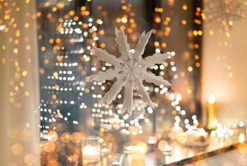 christmas and holidays concept - paper snowflake decoration hanging over garland lights and candles...