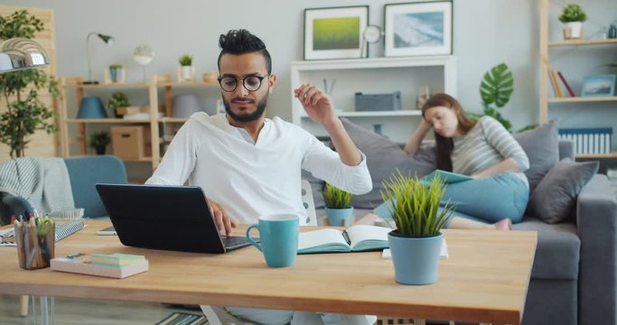 Tired freelancer handsome Arabian man is working with laptop then relaxing stretching arms while Caucasian woman is reading book in background. Family and lifestyle concept.