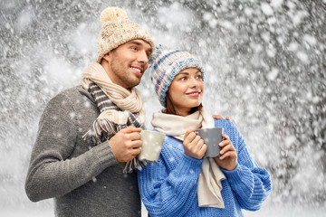 Fototapeta premium people, christmas and love concept - happy romantic couple in knitted hats and scarves with mugs over snow background