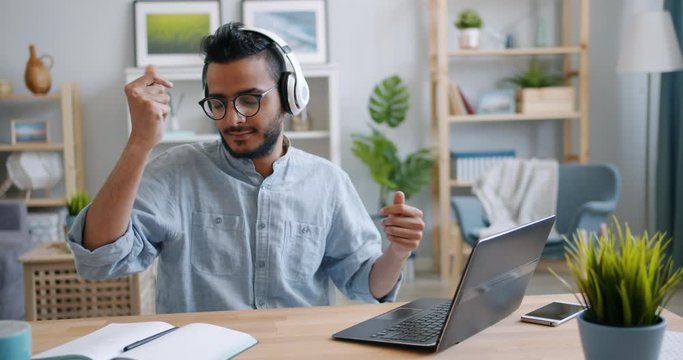Handsome young Arab is enjoying music in headphones and working with laptop at home sitting at table having fun. People, devices and entertainment concept.