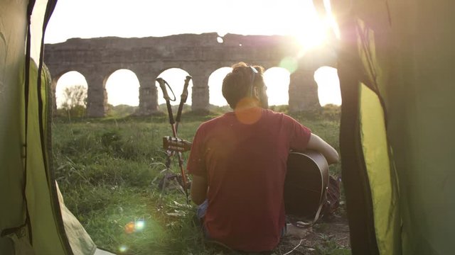 Young man backpacker playing guitar outside camping tent sitting on grass in front of roman aqueduct arches in parco degli acquedotti park ruins in rome at sunset sticks in ground camera shot inside