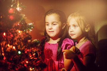 Obraz na płótnie Canvas Two little girls looking at the christmas fireplace near beautiful Christmas tree. Twins in red dresses looking at Santa in fireplace.