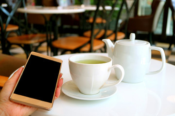One Cup of Hot Green Tea and Teapot on Cafe's Table with Female's Hand Holding a Mobile Phone