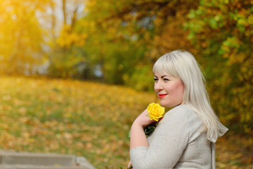 Autumn portrait of a smiling middle-aged blonde woman who stands and poses in the Park on a Sunny day with a yellow rose in her hand. She looks away and smiles. Conceptual photography.