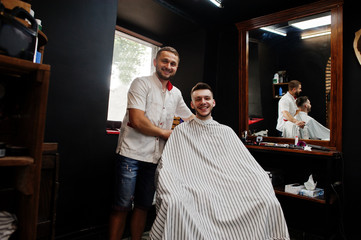 Obraz premium Young bearded man getting haircut by hairdresser while sitting in chair at barbershop. Barber soul.