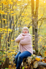 A bright middle-aged woman in a delicate knitted jumper poses on a Sunny autumn day in the forest on logs. Arms crossed over his chest. He looks up dreamily. Woman's size plus xxl. Vertical view