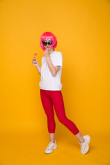 Woman model in the yellow studio with lollipop in a pink wig and bright clothes