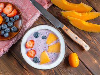 Top view of blueberry smoothie in a coconut bowl with strawberries and melon and kumquat on a red towel with a teaspoon and knife.