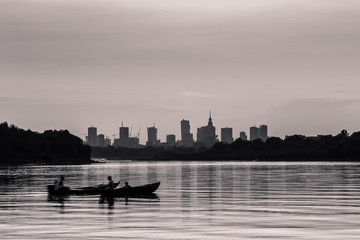 Monochrome Warsaw cityscape with skyscrapers in the downtowan and two boats sailing on the Vistula River during sunset.