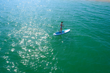 Fototapeta premium Aerial drone birds eye view of young woman exercising sup board in turquoise tropical clear waters