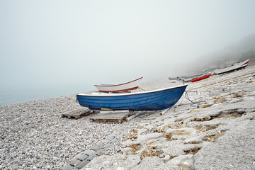 Blue and red fishing boats on a rocky pebble beach in the early morning with foggy mist on background - 287442325