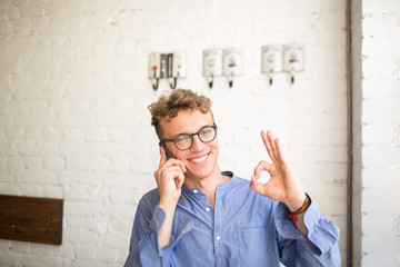 Young smiling businessman showing fingers okey while talking on mobile phone. Happy hipster guy having cell telephone conversation looking in camera while sitting in coffee shop or office space