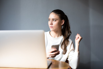Woman copywriter holding mobile phone and dreaming about something during work on laptop computer. Thoughtful female looking away and us cellphone and net-book. Hipster girl waiting call on smartphone
