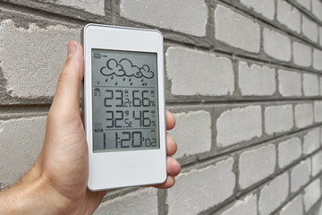 Best personal weather station device with weather conditions inside and outside. A man holds a gadget in his hand against a brick wall.