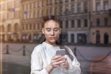 View through coffee shop window with city reflection of a young woman student chatting on mobile phone. Female reading text message on cellphone. Hipster girl received a notification on cell telephone