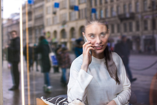 View through coffee shop window with street reflection of a hipster girl having mobile phone conversation with friend while sitting in restaurant. Woman talking on cell telephone during rest in cafe