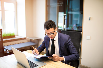 Successful businessman reading information in notepad while sitting at the table with laptop computer in office of financial company. Male proud CEO holding textbook during work on portable netbook - obrazy, fototapety, plakaty