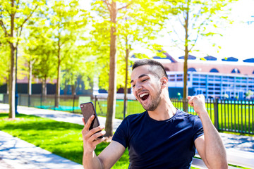 Happy hipster guy received good news in text message on his mobile phone, hand up and said: hooray. Joyful winner man rejoicing his success, holding cellphone. Male having video call via cellular