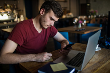 Young serious man writes the information on the sticker from e-mail on mobile phone during webinar on laptop computer. Hipster guy skilled freelancer writing notary during online training course