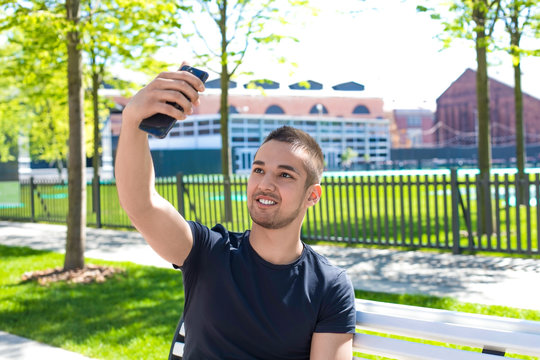 Handsome male making selfie photo on mobile phone camera while sitting in city park in sunny summer day. Smiling man having online video call on smartphone during free time outdoors