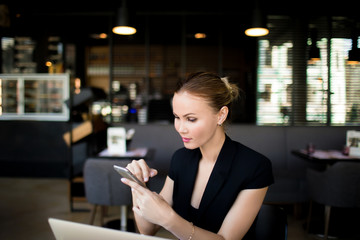 Successful business woman checking e-mail via smartphone after work on laptop computer during coffee break in restaurant. Female entrepreneur typing text on cellphone. Lawyer using apps on mobile