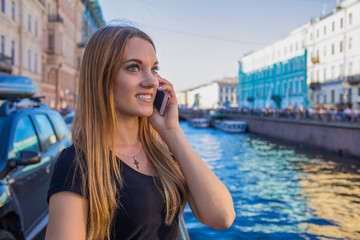 Joyful woman tourist talking via mobile phone while standing near river quay during summer trip. Smiling female with good mood having smartphone conversation during walking outdoors