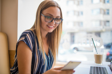 Happy smiling woman freelance social media content writer reading text message on mobile phone during work on laptop computer,sitting in coffee shop during recreation time.Joyful hipster girl chatting