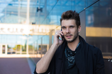 Young handsome casual dressed hipster guy talking via mobile phone while standing outdoors near business centre background with copy space for advertising content. Bearded man phoning via cellphone