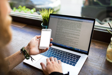 Hipster guy reading text message on mobile phone with empty screen with copy space for promotional content during online learning on laptop computer sitting in coworking space. Man using cellphone