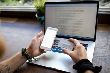Man holding mobile phone with empty screen with copy space and credit card doing online order in internet while sitting with laptop computer in coffee shop. Male online banking via cellphone