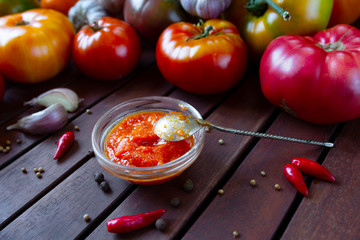 Homemade Adjika tomato sauce in bowl with fresh ingredients - Tomatoes, chilli pepper, garlic on wooden table.