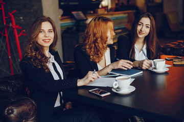 three beautiful elegant girls in white shirts and black jackets sit at the restaurant at the table and use notebooks and pens, they also drink coffee