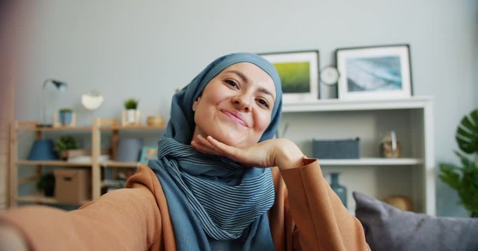 POV slow motion of happy Middle Eastern woman in hijab taking selfie at home posing looking at camera having fun. People, lifestyle and modern devices concept.