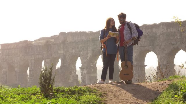 Young lovely couple backpackers tourists reading map pointing directions roman aqueduct arches in parco degli acquedotti park ruins in rome on romantic misty sunrise with guitar and sleeping bag slow