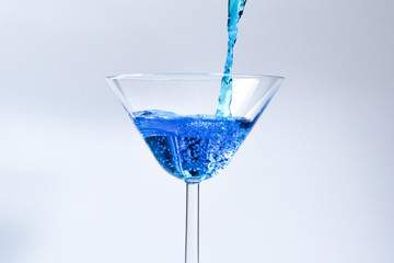 Cocktail with blue liquid in glass. Glass with blue water pouring with liquid with splashes and drops. Martini glass filling with alcohol with splashes on white background. Refreshing drink concept.