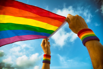 Hands with rainbow color wristbands waving LGBTQI gay pride flag in the wind against a vibrant sunny blue sky