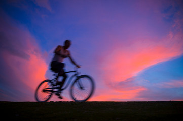 Naklejka na ściany i meble Defocus abstract sunset view of unrecognizable silhouette of a man cycling in motion blur against colorful sky