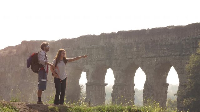 Young lovely couple backpackers tourists reading map pointing directions roman aqueduct arches in parco degli acquedotti park ruins in rome on romantic misty sunrise with guitar and sleeping bag slow