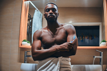 Attractive young man in towel is brushing his teeth in the bathroom while sitting on the table.