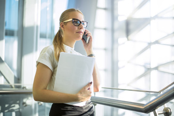 Confident woman in office wear and classic glasses and paper documents in hand having cell telephone conversation while standing in contemporary enterprise interior.Female banker phoning via cellphone