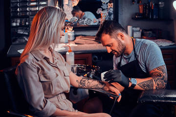 Dilligent focused tattoo artist is creating new tattoo on young woman's hand at tatoo studio. - obrazy, fototapety, plakaty