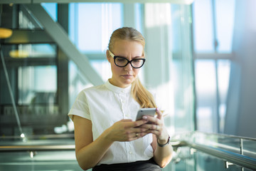 Frustrated woman manager in official wear and glasses received on mobile phone e-mail with bad news, standing in enterprise interior. Upset female CEO reading problem notification on cell telephone