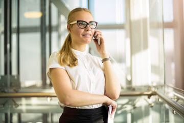 Fototapeta premium Smiling woman in official wear successful government worker talking via smartphone while standing in modern interior during break at job. Joyful female in glasses skilled entrepreneur phoning