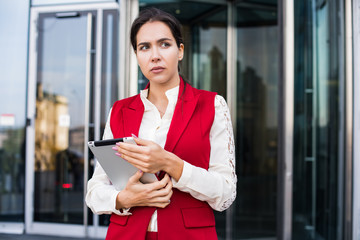 Fototapeta na wymiar Pensive woman manager thinking about work issue while standing with portable touch pad near entrance to company while waiting International partners. Thoughtful serious female office worker outdoors