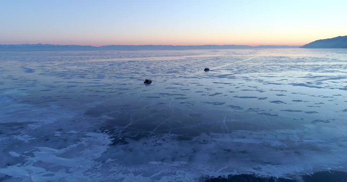 Aerial: Amphibious vehicles floating on frozen Lake Baikal against blue and orange sky