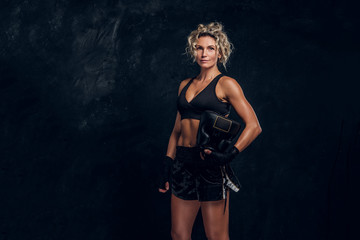Fototapeta na wymiar Experienced female boxer is posing for photographer at dark photo studio with equipment in her hands.