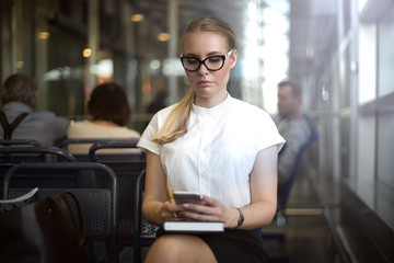 Serious woman office worker checking e-mail on mobile phone while sitting in waiting room. Female manager in glasses using apps on cell telephone, sitting in airport terminal before business trip